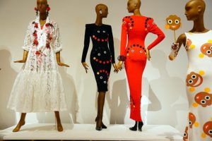In this installation photo, 4 mannequins pose in garments designed by Patrick Kelly. The mannequin on the left wears a drapey white lace midi dress ornamented with red roses. Two mannequins wear tight fitting black dresses with sharp shoulders embroidered with the image of a golliwog, a racist african american cariacture that looks similar to a smiley face emoji. One dress is black and one dress is red. The mannequin on the white wears a tight fitting white sheath dress also decorated with the golliwog. In her hand she carries a mask of the golliwog.