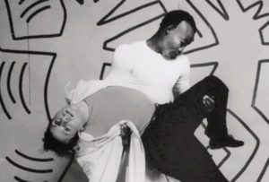 A black and white photo of two dancers in front of a Keith Haring designed set. One dancer holds another with one arm under their back and the other under their knee.