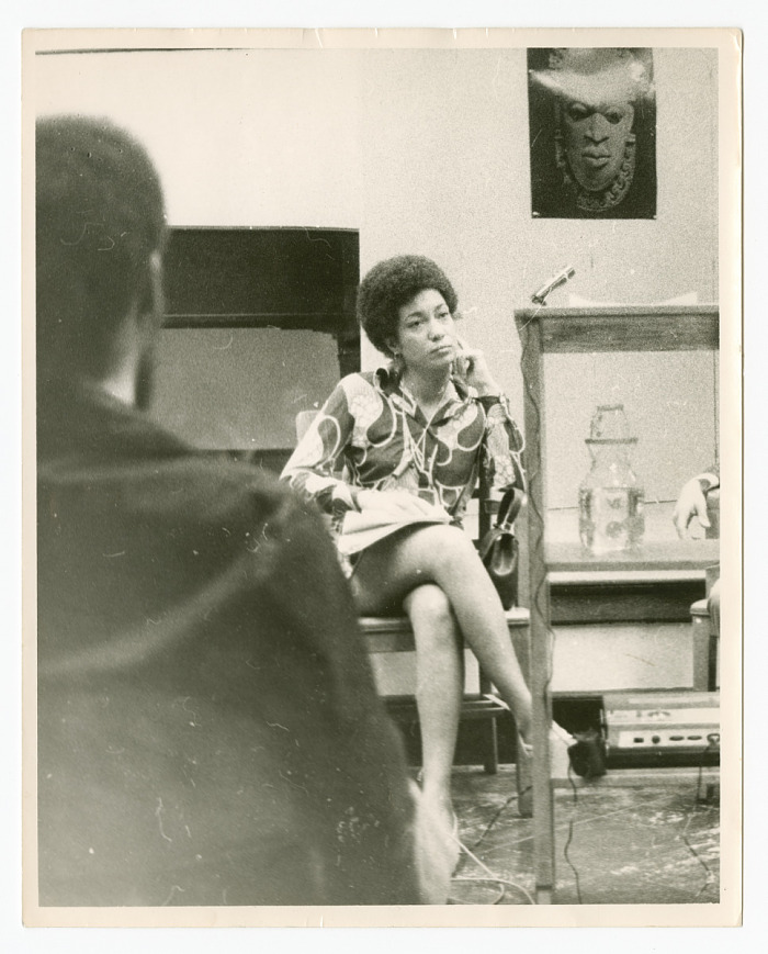A black and white photograph of poet June Jordan. Jordan sits in a chair with her proper left arm resting against her cheek. An unidentified person sits with their back towards the camera. The back of the photograph has an inscription identifying the photograph’s subject.