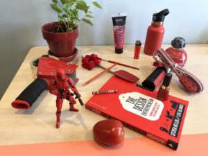 A collection of red objects arranged on a desk. The objects include toys, books, an apple, spatula, and lotion bottle.