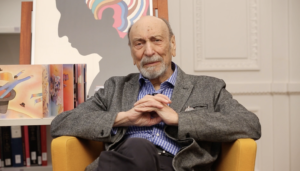 Image of Milton Glaser, a older white man with thin hair on the sides of his head. He sits in a bright yellow chair with his hands clasped in front of him.