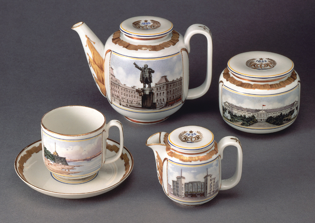 Image features a ceramic tea service composed of a hot water pot, sugar bowl, creamer, and cup and saucer. Each simple cylindrical vessel is decorated on one side with a colorful urban scene featuring a Soviet monument or state building in Leningrad. The opposite side of each piece is decorated with a gilded image of a historical site in Leningrad. The pieces are further embellished with classically-inspired gilded ribbons and marshal trophies at the edges, spouts, and loop handles. Please scroll down to read the blog post about this service.