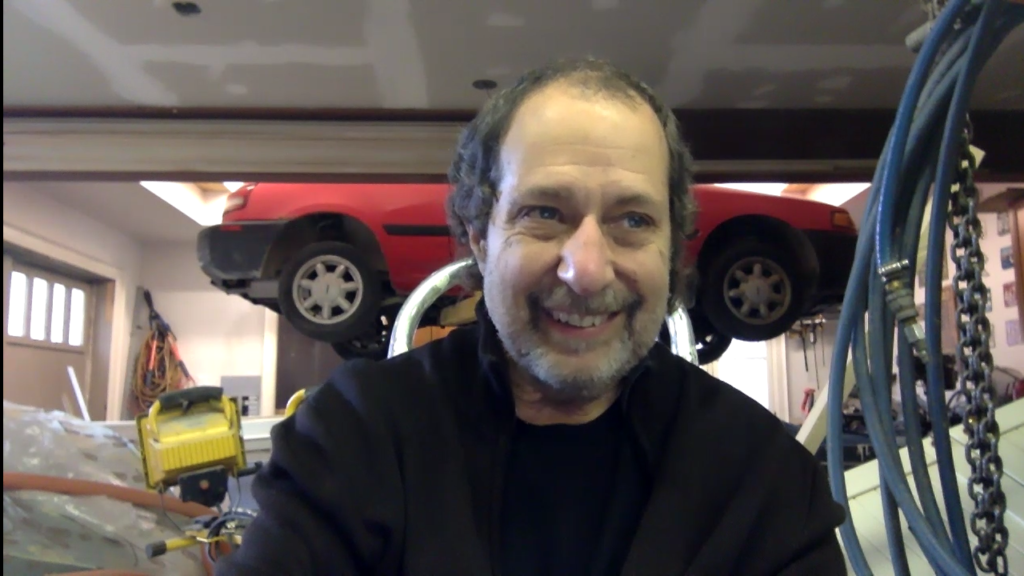 Rick Valicenti, screenshot from a Zoom call. Rick is smiling and wearing a dark sweater. Behind him, a red car is suspended in the air for repairs.