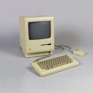 Beige rectangular computer (a) with glass screen above computer with glass screen and floppy disc drive, plastic keyboard and mouse with connecting wires.