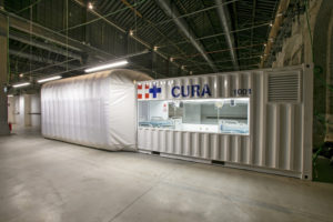 White temporary structure with CURA written on the side and medical facilities visible inside housed in a larger warehouse space