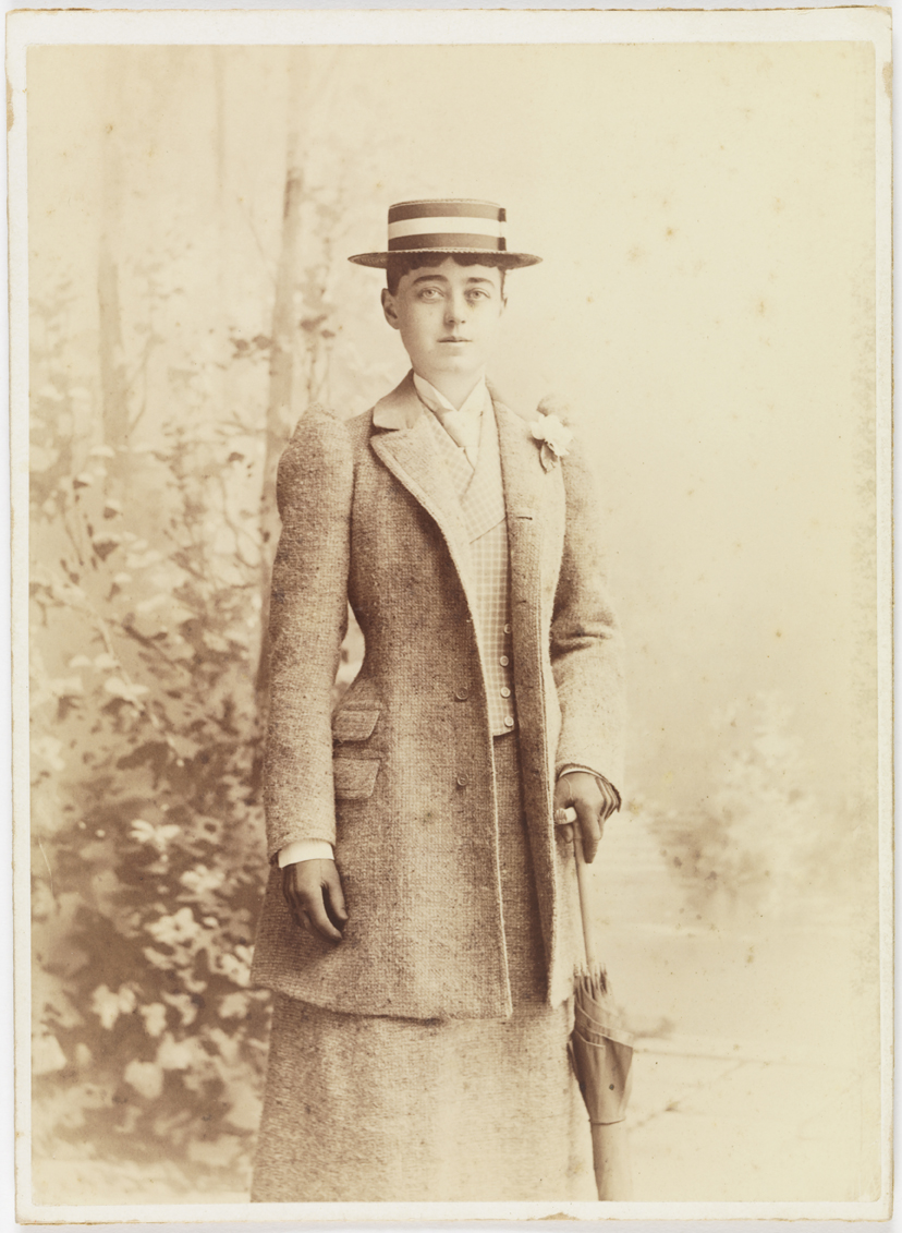 Full-body portrait of Sarah Hewitt wearing coat, vest, skirt, and hat. She wears a flower on her lapel and holds an upturned umbrella.