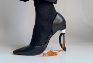 A close-up profile view of a black socked right foot underneath the edge of a black pant cuff, the foot is wearing a black stiletto-style shoe, the high heel is replaced with a vegetable peeler, ribbons of orange carrot peels trail from peeler into a small pile on the white floor.