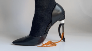 A close-up profile view of a black socked right foot wearing a black stiletto-style shoe, the high heel is replaced with a vegetable peeler, ribbons of orange carrot peels trail from peeler into a small pile on the white floor.