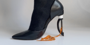 A close-up profile view of a black socked right foot wearing a black stiletto-style shoe, the high heel is replaced with a vegetable peeler, ribbons of orange carrot peels trail from peeler into a small pile on the white floor.