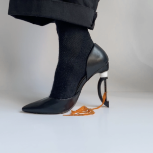 A close-up profile view of a black socked right foot underneath a black pant cuff, the foot is wearing a black stiletto-style shoe, the high heel is replaced with a vegetable peeler, ribbons of orange carrot peels trail from peeler into a small pile on the white floor.