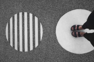 Aerial photograph of two silver circles over a black and white speckled floor. The left circle is sliced by vertical stripes reveling the floor below. On the right, seen from thighs down, a person stands facing inward atop the circle in black sandals and pants.