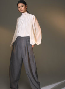 An Asian woman models long tapered grey slacks with a flowing white blouse and gold hoop earrings. The model stands with her hands in her pockets looking directly at the camera.