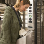 A blonde woman wearing a green knit sweater demonstrates using a 3D printed white accessory. The accessory hangs from her neck is being used to to swipe a NYC metrocard in a subway station.