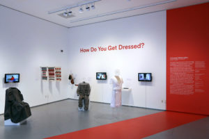 An exhibition showcasing the application of interactive textiles in wearable clothing or accessories. On the white wall behind the clothing in red letters reads [How Do You Get Dressed]?