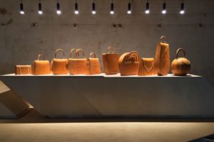 A photograph of ten orange handbags lined up on a concrete table. Each handbag is made of the same material but varies in shape and size.