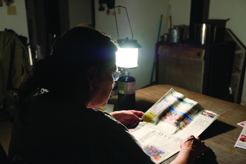 A photograph of a woman seated near a kerosene lamp and reading a newspaper. She is photographed from behind, over her shoulder, as the lamp lights her reading. The scene is situated in a dimly lit interior.