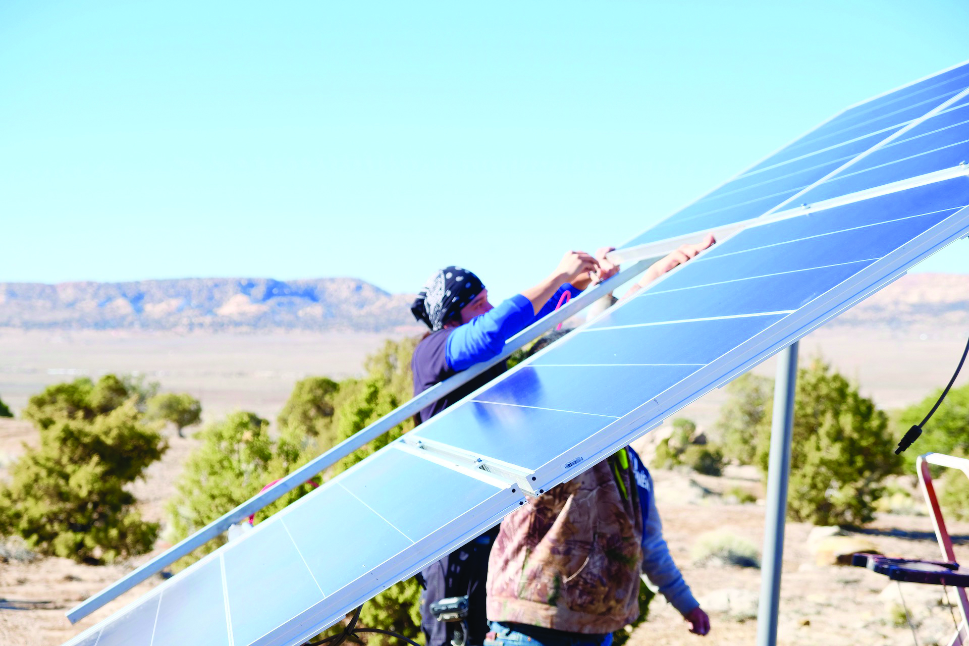 A photograph of two individuals working on a solar panel. The solar panel is seen from the side, dissecting the image from lower left to top right. The two individuals are behind and largely obscured by the panel. In the background is natural imagery of the Southwest of the United States.