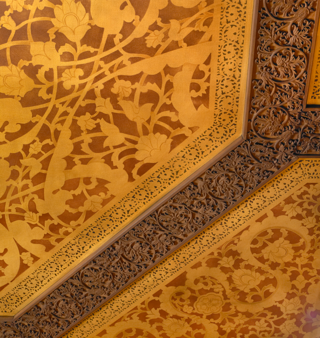 Photo of intricate floral details. Carved, and painted vines with flowers fill the expansive geometric zones of the gold and brown ceiling
