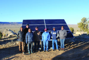 A photograph of seven people standing, smiling and posed, before a solar panel. In the background is natural imagery of the Southwest of the United States.