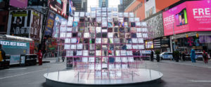 A geometric sculpture comprised of mirrors that make a heart shape. The mirrors reflect the lights and sky of Times Square.