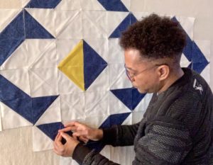 William Daniels, pictured on the right side of the image, works on a blue and white quilt pinned to a wall. William wears a grey shirt and glasses, and is pinning a blue triangular piece of fabric to the outer edge of the quilt. At the quilt's center, a yellow and blue triangle fit together to create a diamond shape.
