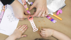 This image shows a parent helping a child add a pink sticker to a watch. Around them you can see a drawing, foil, markers and paper.