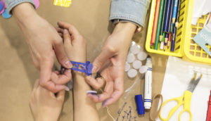 This image shows a parent helping a child attach a purple accessory to their wrist. Next to them, there is a blue marker, scissors with a yellow handle, and a yellow bin with colored markers.