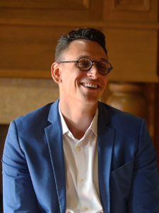 A photograph of a light-skinned man with short dark hair and glasses wearing a blue suit jacket white shirt and grey pants sitting on sofa in a wood-paneled study.