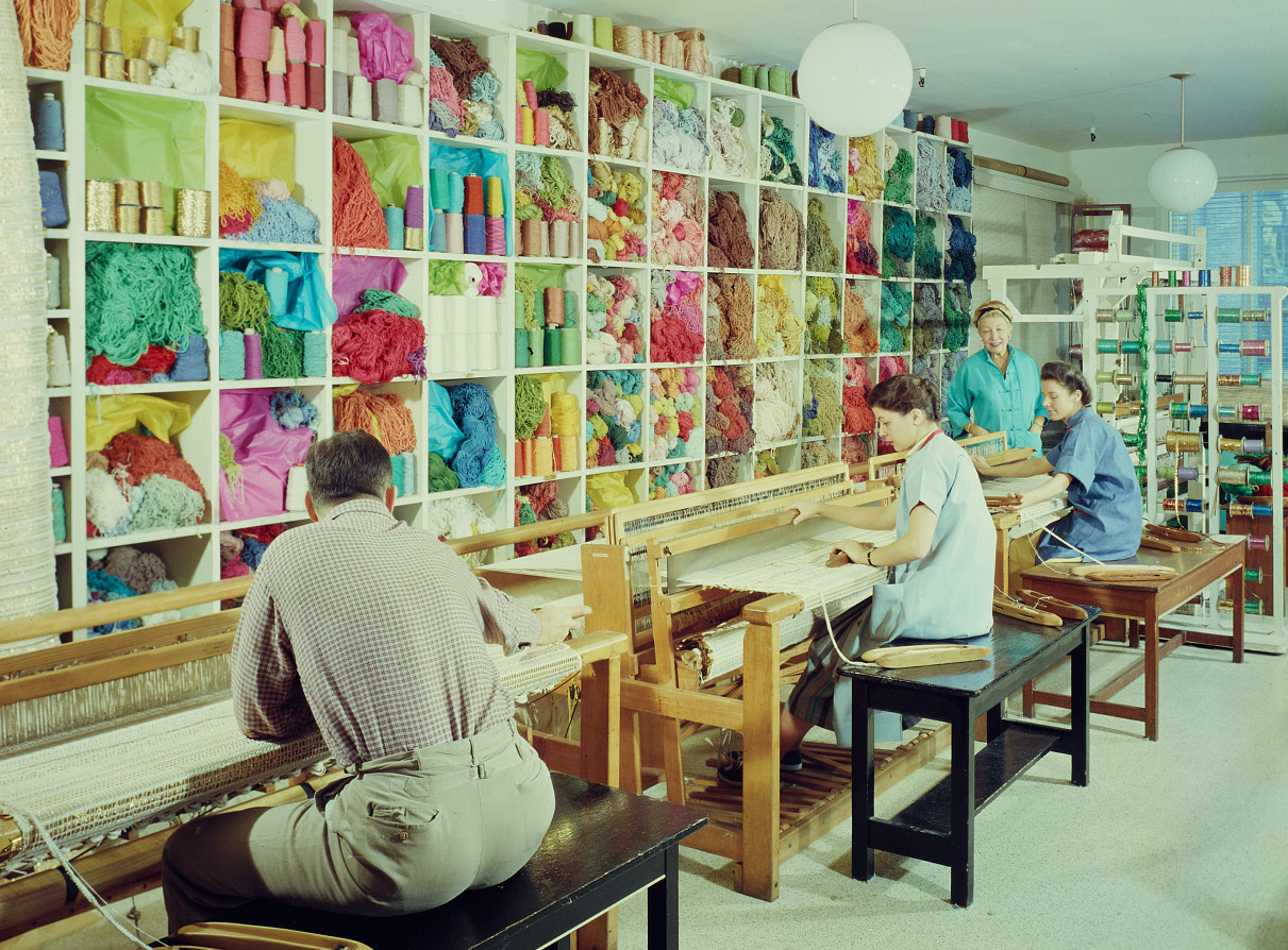 Three people sit working at large textile looms in front of a high wall of shelves filled to the brim with vibrant and colorful yarn and other kinds of thread; a fourth person stands and watches their work.