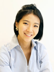 A headshot photograph of a medium-skinned Asian American woman with short black hair wearing a pale blue shirt and a gold hoop earring in her right ear