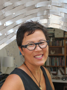 A headshot photograph of a Korean-American short haired woman with geometric glasses wearing a grey V-neck top and a black string necklace with a pearl on the end, behind her a silver-plated arch, a tiered bookshelf and a white shaded lamp on a desk.