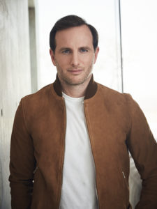 A headshot photograph of a light-skinned man with short dark hair and brown eyes wearing a tan leather jacket and a white t-shirt, behind him is a window and a light grey wall.