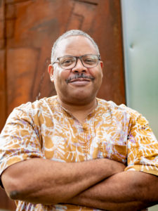 A photograph of a dark-skinned man wearing glasses and an orange and white patterned shirt with his arms folded across his chest, behind him is a wooden wall.