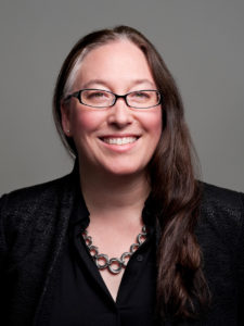 A headshot photograph of a light-skinned woman with long dark hair and thin-framed rectangular black glasses wearing a black shirt and a thick grey chainmail necklace.