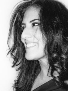 A black and white headshot photograph of a light-skinned smiling woman facing to our left with long dark hair that curls at the end.