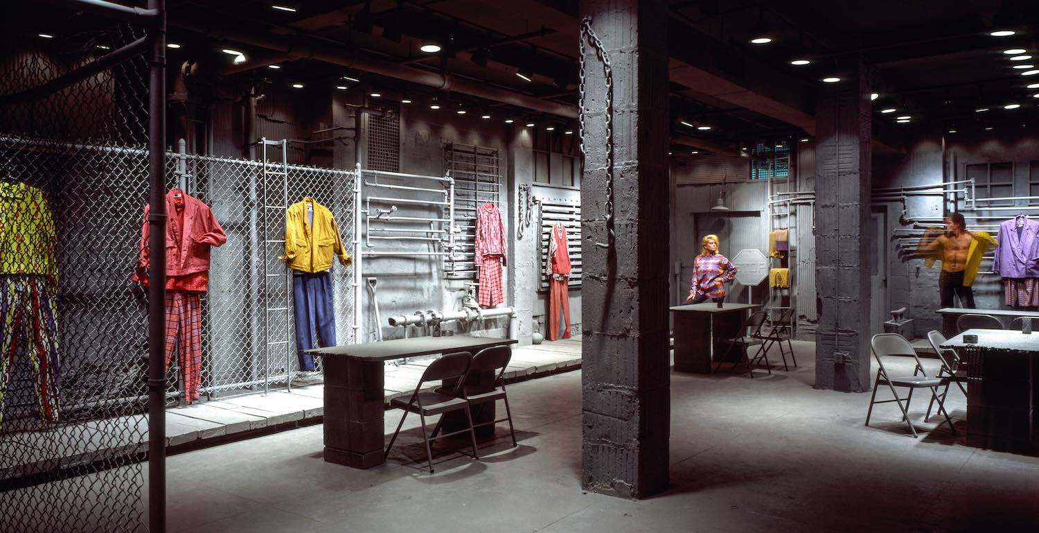 A WilliWear showroom. In an urban / industrial gray warehouse space, garments are suspended not from clothing racks but from chain link fence that lines the wall. The garments are simple blouses and pants. In the background, two models, one woman and one man, look at each other like they're sizing each other up