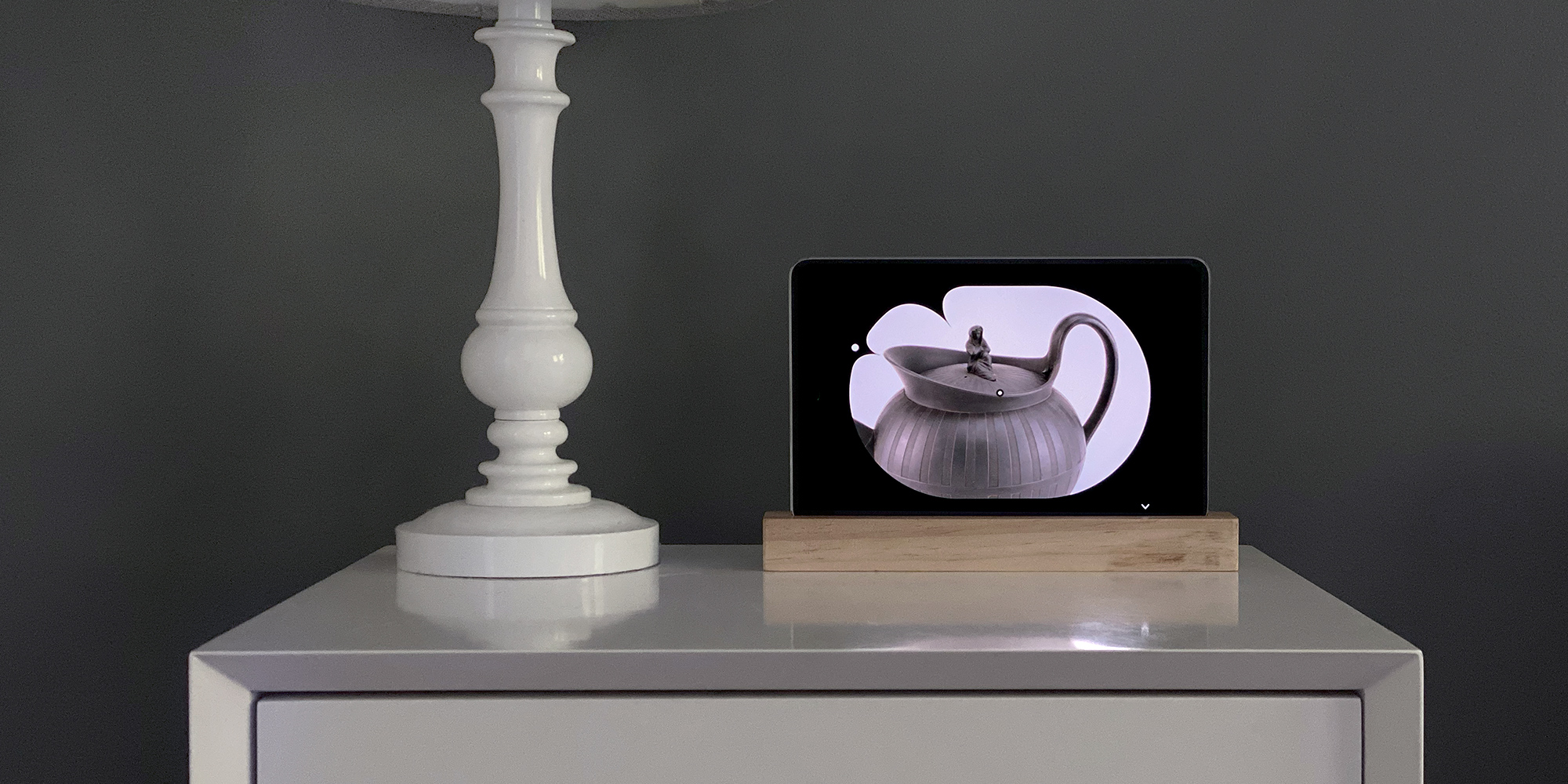 Art Clock interface being visualized on a tablet that is sitting in a stand on top of a gray cabinet next to a decorative white candlestick. The interface itself shows a stylized clock at 11:50 with the clock hands aligning to different parts of the teapot it displays.