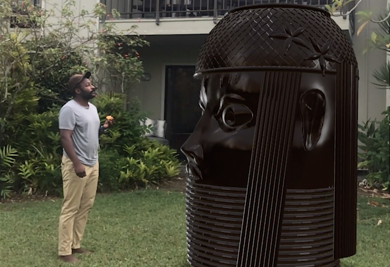 A Black person wearing a baseball cap, grey t-shirt and khaki pants standing on a lawn and gazing at a larger than life-size augmented reality rendering of a bronze King's Head sculpture from the Kingdom of Benin. The background is the landscaped exterior of an apartment building.