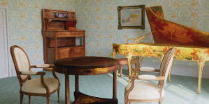 Augmented reality room meant to evoke the Belle Epoque in Paris in 1912. The room is furnished with wall coverings, textiles and objects of the period, including an Art Nouveau wallpaper in mint green with orange accents, a wooden hutch, a gold harpsichord with paintings along the side, a wooden table with gold accents, and two wooden chairs upholstered in white fabric. Light streams into the room from the right.