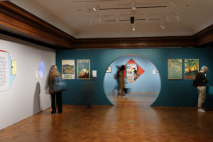 View of gallery wall painted blue with large circular opening, flanked on either side by vertical posters and visitors looking at artwork. Circular opening provides sightline to other parts of the exhibition and more visitors within the museum.