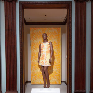 Gallery view of exhibition showing mannequin wearing floral shift dress in bright orange and pale yellow, set against large floral textile in similar colors.