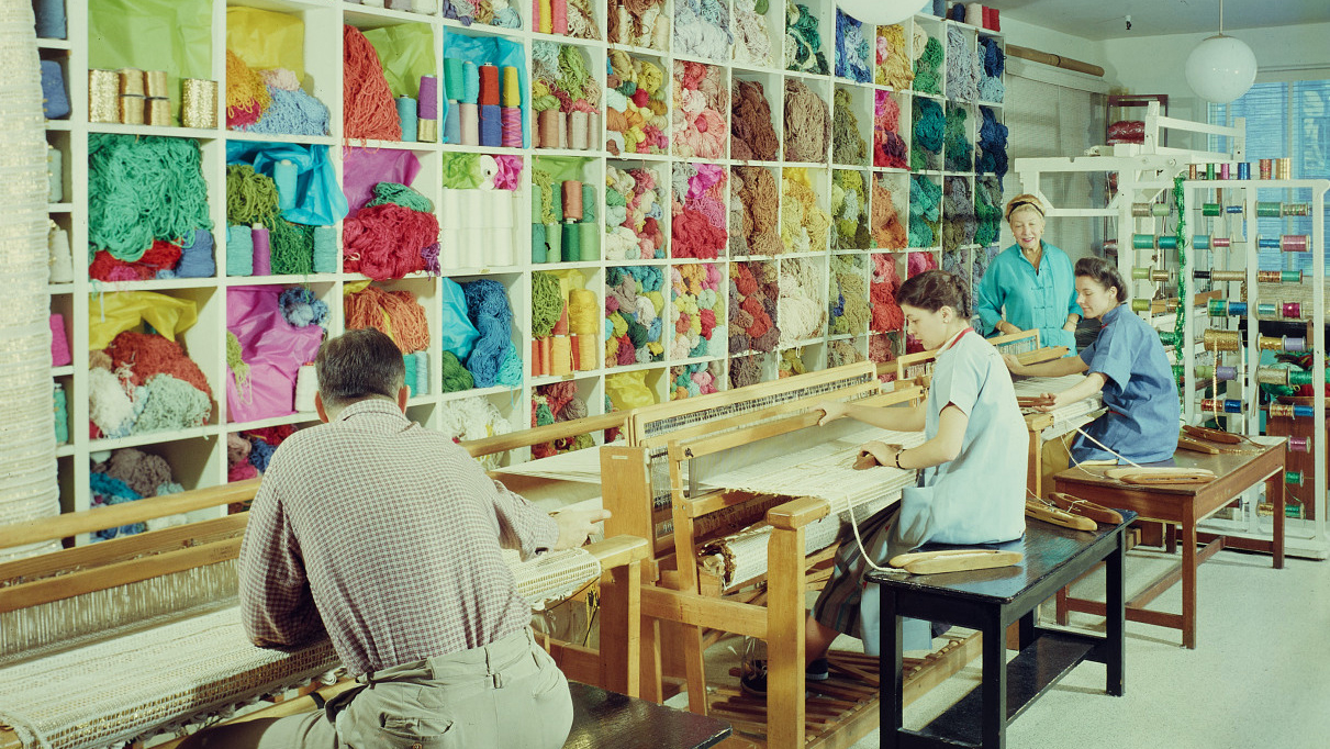 Three people sit working at large textile looms in front of a high wall of shelves filled to the brim with vibrant and colorful yarn and other kinds of thread; a fourth person stands and watches their work.