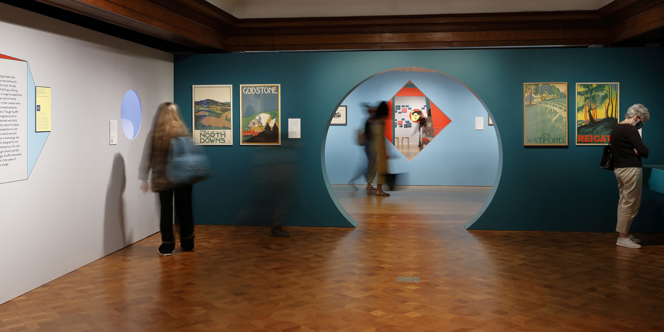 View of gallery wall painted blue with large circular opening, flanked on either side by vertical posters and visitors looking at artwork. Circular opening provides sightline to other parts of the exhibition and more visitors within the museum.