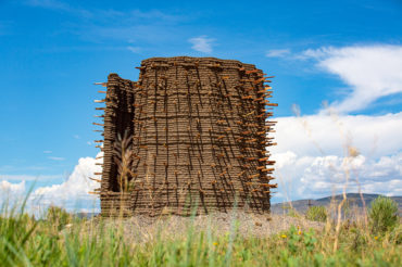 Large circular, adobe structure with sticks protruding from the walls, set against distant mountains and crystal blue sky.