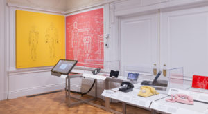 A waist-high shelf holds equipment, including historic telephones and a touchpad display. In the corner, yellow and pink graphics show diagrams of the human body.