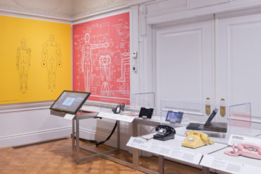 A waist-high shelf holds equipment, including historic telephones and a touchpad display. In the corner, yellow and pink graphics show diagrams of the human body.