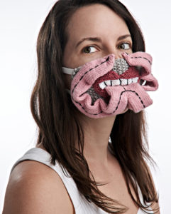 A woman wears a knitted face mask depicted a cartoonish mouth full of teeth and a large, sculptural bifurcated pink tongue that forms ribbonlike strips that end in points at the wearer's nose