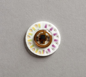A plastic white disc with round brown circuitry at its center and indentions around the circumference in purple and yellow