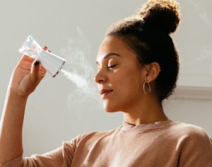 A dark-skinned woman closes her eyes and inhales from a white hourglass-shaped mister held several inches from her face, from which stems a thin cloud of white steam