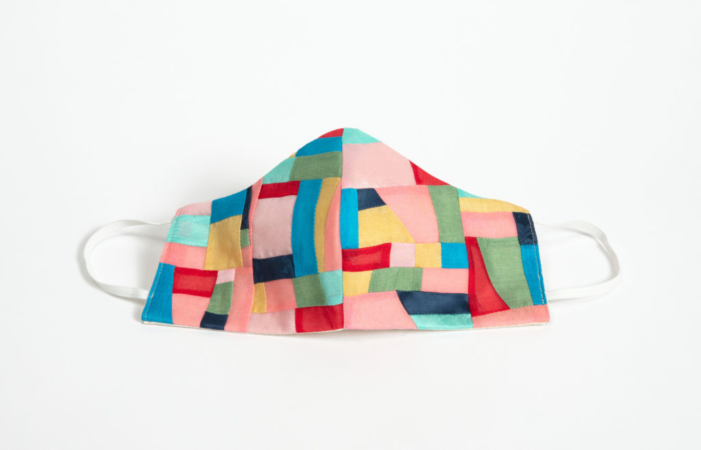 A colorful face mask made up of irregular geometric silk shapes in shades of pink, blue, yellow, and green